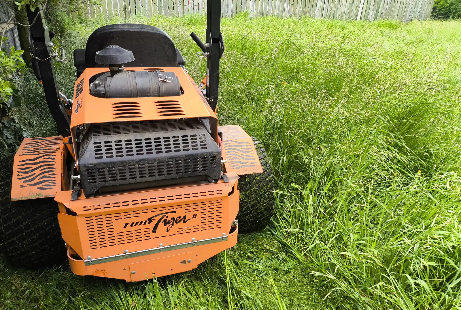 Orange Turf Tiger ride-on mower cutting a strip through long, overgrown grass beside a timber fence